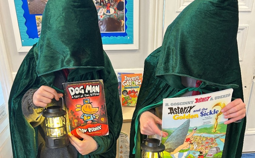 two pupils dressed up in green cloaks holding lamps and books