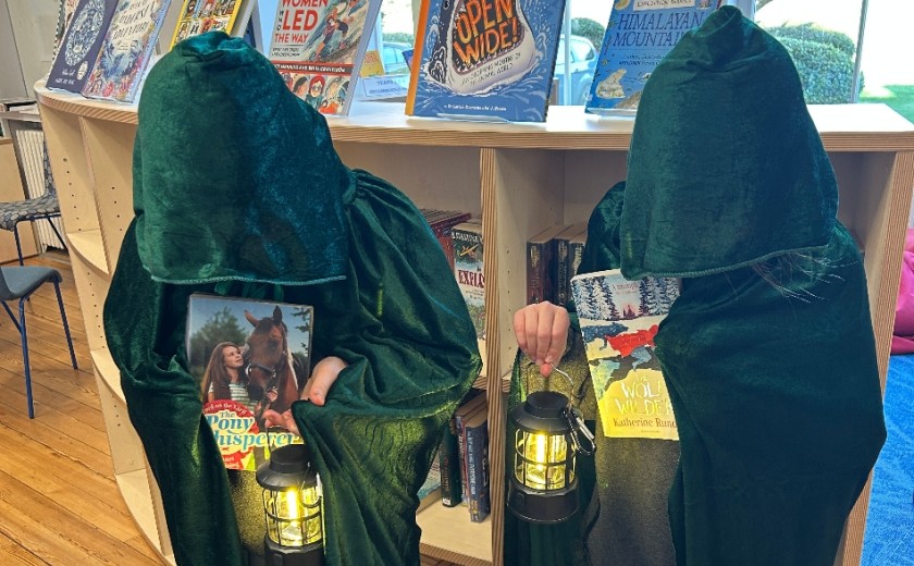 two girls wearing green cloaks holding lamps and books